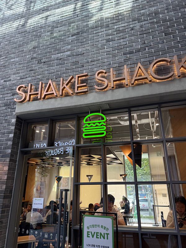 Menu board showcasing Shakeshack's offerings including burgers, fries, shakes, and beverages against an urban exterior backdrop.