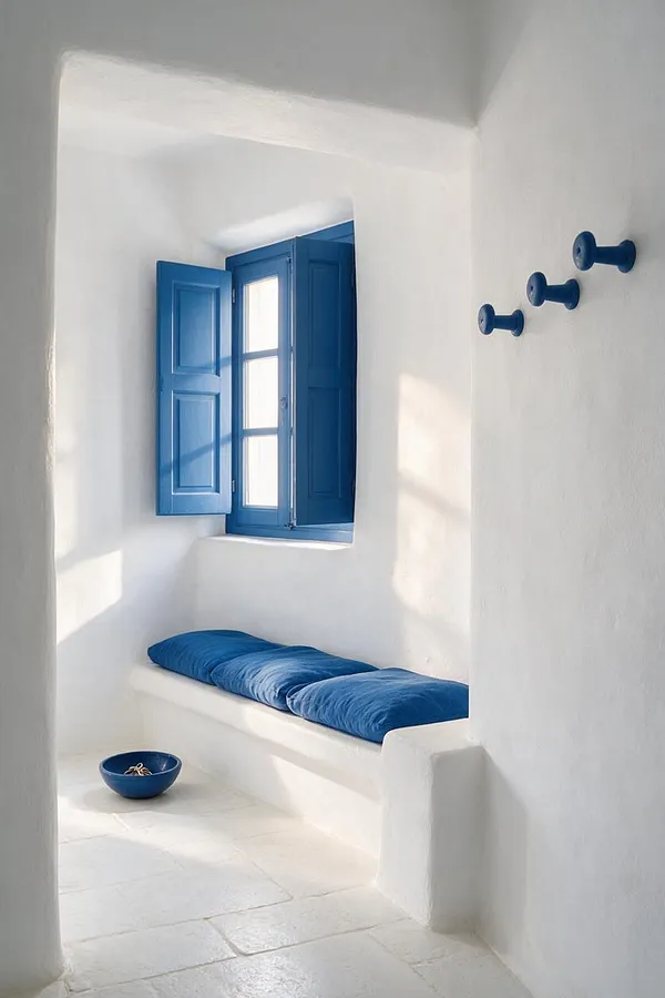 A narrow Mediterranean-style entryway with whitewashed plaster walls, a blue shuttered window, and a built-in bench topped with blue cushions. Three blue wall pegs and a small blue bowl on the pale stone floor complete the serene, coastal look.