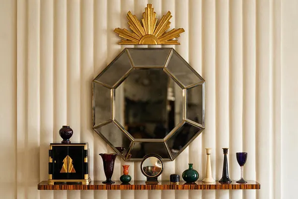 An Art Deco-inspired wall display features an octagonal mirrored frame with metallic edging and a gold sunburst ornament centered above it. Below, a narrow striped wood shelf holds small decorative glass vessels, a black lacquer box with gold detailing, and a few candlestick-like objects against soft cream drapery.