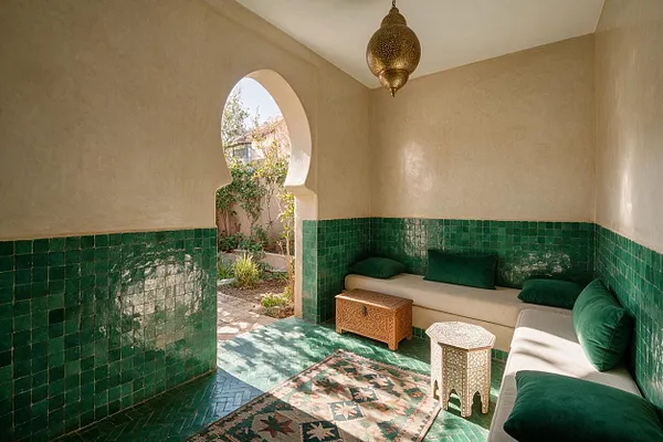 A Moroccan-style sunroom with glossy green tile wainscoting, warm plaster walls, and an arched doorway opening to a garden. Built-in seating with cream cushions and deep green pillows lines the walls, with carved wood and white Moroccan-style side tables and a patterned rug on the floor.