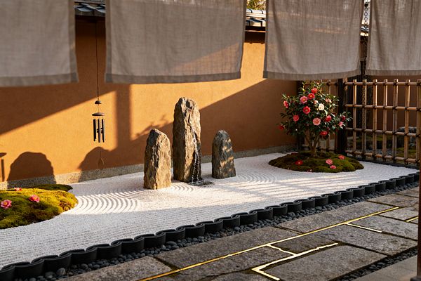 A compact Japanese Zen patio features three upright basalt stones, one spilling a thin sheet of water, centered in meticulously raked white gravel with concentric circles. Moss mounds with pink camellia blossoms flank the stones, bordered by a bamboo lattice and scalloped edging, while an ochre plaster wall, hanging linen noren, a dark metal wind chime, river pebbles, and stone pavers with brass inlay catch the warm afternoon light.
