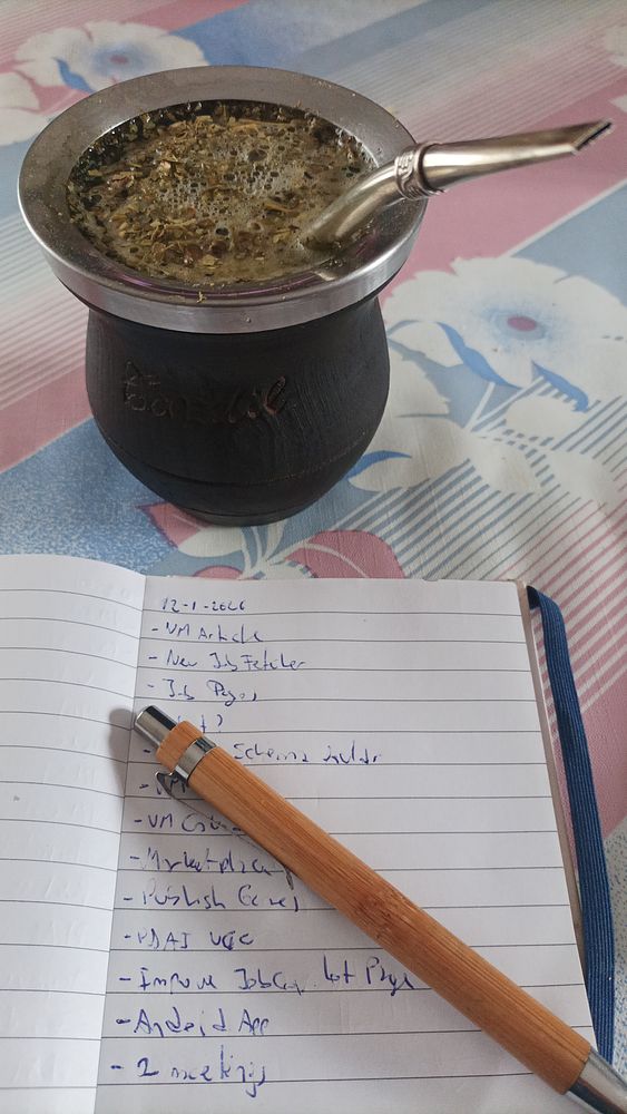 A person holding a traditional mate cup with a metal straw inside, accompanied by a notebook and a pen on a wooden table.