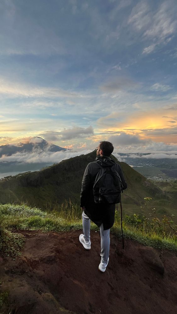 A hiker stands on the edge of a mountain, gazing at a colorful sunrise over the landscape.
