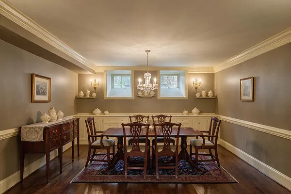 A traditional basement dining room with taupe walls, white crown molding and wainscoting, a dark wood dining table, six carved wooden chairs, and a patterned area rug. A crystal chandelier hangs centered over the table, with built-in cabinetry, decorative porcelain pieces, framed artwork, and small windows at the back wall.