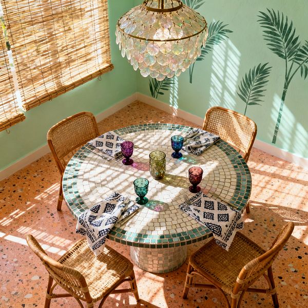 A sunlit Bohemian dining nook centers on a round mosaic-tiled pedestal table set with multicolored glass goblets and indigo patterned napkins, surrounded by four rattan chairs. Mint green walls painted with palm fronds, an iridescent capiz shell chandelier with brass details, warm terracotta terrazzo flooring, and woven bamboo window shades casting striped shadows create a tropical, textural atmosphere.