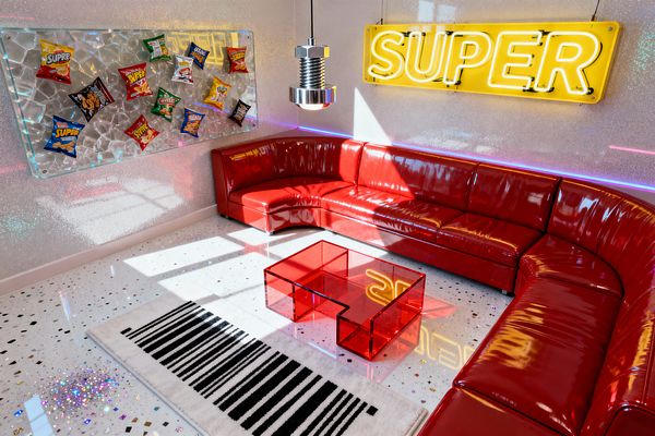 Sunlit living room with a glossy, curved red sectional wraps around a translucent red acrylic coffee table atop a white floor embedded with confetti-like chips and a black-and-white barcode-stripe rug. Iridescent glitter walls host a large yellow neon sign reading "SUPER," purple-blue LED strip lighting, a polished chrome pendant shaped like an oversized bolt, and a wall art panel of snack bags mounted over cracked glass, creating a playful, high-energy Pop Art scene.