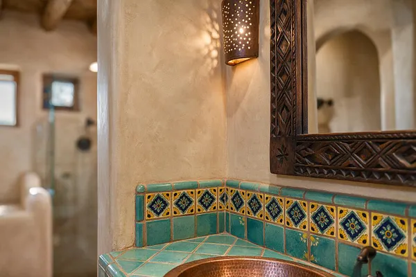 A Southwestern bathroom vignette with a turquoise tiled countertop and backsplash, accented by yellow patterned Spanish-style tiles. A copper sink sits below a carved dark wood mirror, and a perforated metal wall sconce casts warm patterned light on the textured plaster wall.