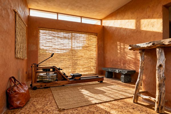 A serene home gym with earthen terracotta plaster walls and cork flooring holds a walnut-wood water rowing machine on a jute rug before floor-to-ceiling bamboo blinds. Sunlight from a clerestory window casts warm patterns across a rough charcoal stone bench, a rustic log console, a woven straw wall hanging, and a leather duffel, highlighting natural textures and a minimal layout with generous open floor space.