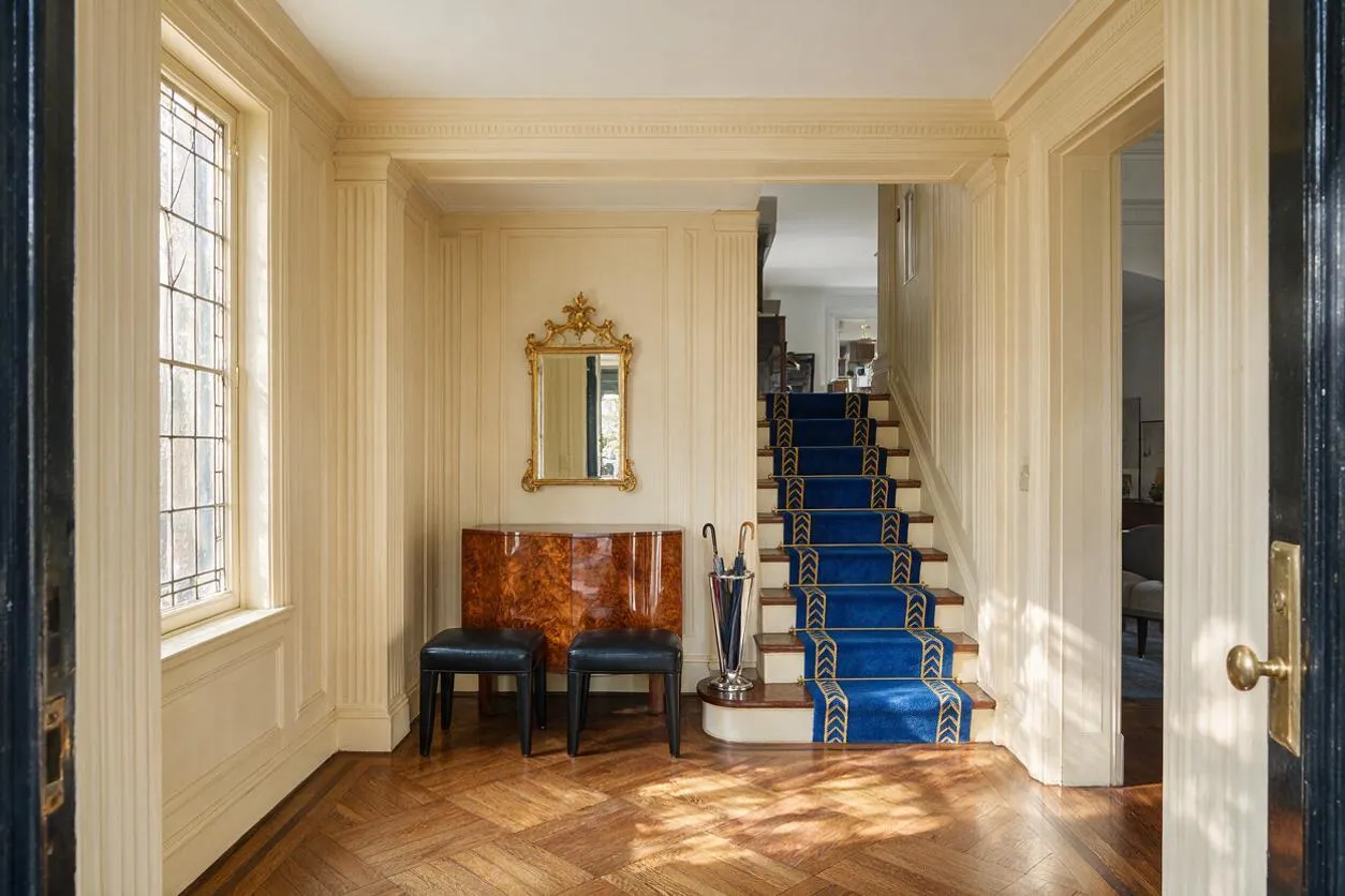 An Art Deco entryway with cream paneled walls, a herringbone wood floor, and a staircase covered in a vivid blue runner with gold geometric trim. A small lacquered console, black stools, a gold-framed mirror, and a chrome umbrella stand create a refined, classic look.