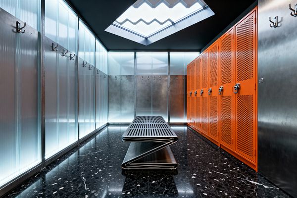 An industrial mudroom with bright orange perforated metal lockers lines the right side, while ribbed translucent panels with rows of metal hooks span the left. A slatted bench made from folded stainless steel sits centered on a glossy black terrazzo floor. Cool light from LED strips glows behind the panels, and a geometric skylight above adds daylight. Brushed metal wall sections and heavy-duty hardware complete the utilitarian, high-performance look.