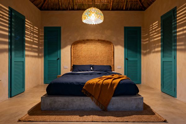 A tropical bedroom centers on a concrete platform bed with deep navy bedding and a tall woven rattan cane headboard, flanked by four teal louvered doors. Warm sand-plaster walls, a thatched ceiling with bamboo rafters, a dome mosaic-glass pendant, a mustard throw, and a natural jute rug complete the scene. The pendant casts dappled shadows across the walls and floor, creating a cozy, resort-like atmosphere focused on natural textures and saturated coastal color.