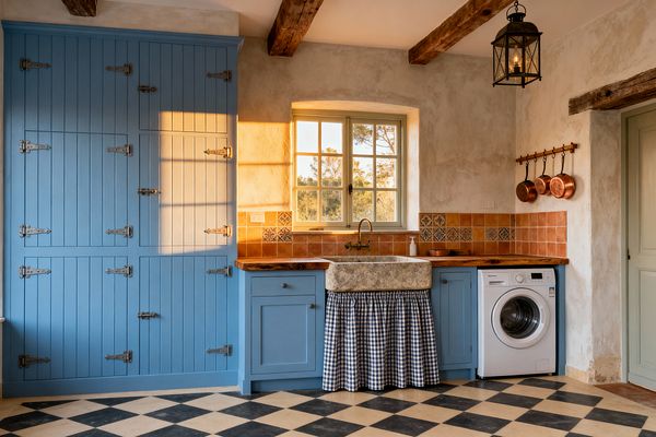 A French Country laundry room features powder-blue beadboard cabinets with a tall larder wall, a carved stone farmhouse sink skirted in blue-and-white gingham, and a terracotta tile backsplash with patterned accent tiles. Reclaimed wood countertops, exposed ceiling beams, a black metal lantern pendant, and a wall-mounted brass faucet add rustic character. A white front-loading washer sits under the counter, with copper pans hung on a rail and a sage-trim window glowing with warm light.