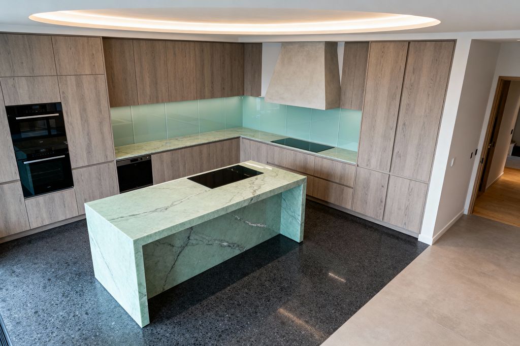 A contemporary U-shaped kitchen with a mint green stone waterfall island holding an induction cooktop. Flat-panel light oak cabinets span two walls with matching stone countertops and a pale aqua back-painted glass backsplash. Black built-in double ovens and a concealed dishwasher sit flush in the cabinetry. A light-gray plaster hood anchors a second induction zone. Dark terrazzo flooring defines the work area beside lighter tile, under a circular cove of warm recessed lighting.