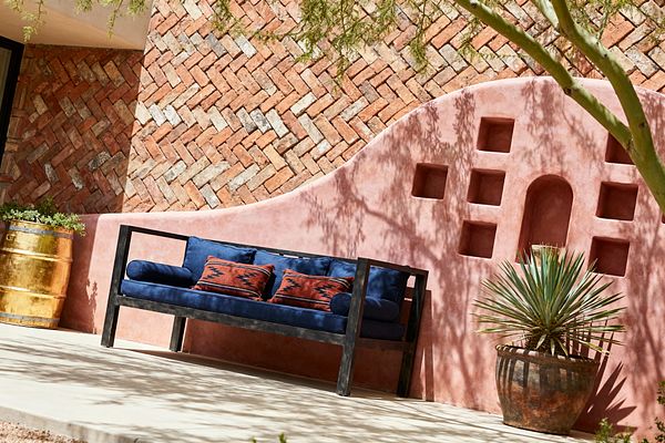 Sunlit Southwestern patio featuring a curved pink adobe stucco wall with square and arched niches in relief. In front sits a navy outdoor bench with a black metal frame, layered with indigo cushions and rust kilim pillows. Behind, a herringbone clay brick wall adds texture. A patinated brass barrel planter with succulents sits left, and a terracotta pot with a spiky agave sits right, on a pale concrete floor dappled by tree shadows.