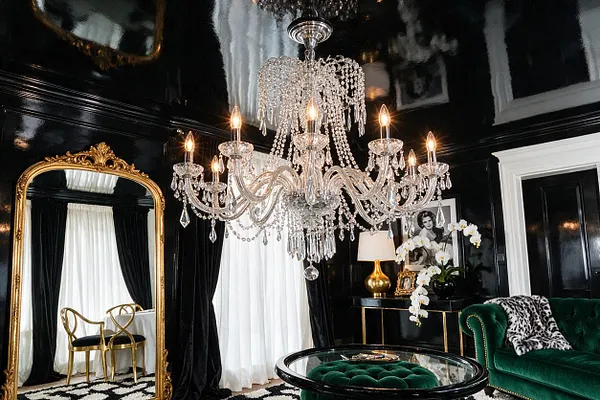 A glamorous Hollywood Regency room with glossy black walls and ceiling, a large ornate gold-framed mirror, and a crystal chandelier centered overhead. Deep green velvet seating, gold accents, sheer white curtains, and a black-and-white patterned rug complete the dramatic, luxurious look.
