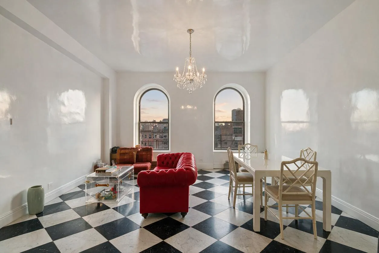 A bright studio apartment with high-gloss white walls, a black-and-white diamond checkered floor, and two tall arched windows framing a city view. A crystal chandelier hangs in the center, while a red velvet tufted sofa and a clear acrylic coffee table sit beside a cream dining table with matching chairs.