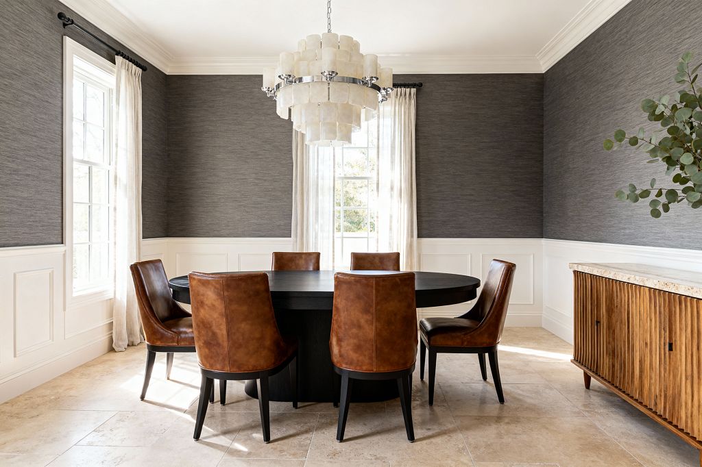 A transitional dining room features a round black pedestal table with six cognac leather chairs on a beige stone tile floor. The walls combine charcoal gray textured wallpaper above white paneled wainscoting, with two tall windows dressed in off-white linen curtains. A tiered alabaster chandelier with polished nickel hardware hangs over the table. At right, a fluted wood sideboard with a pale stone top holds a vase of eucalyptus.
