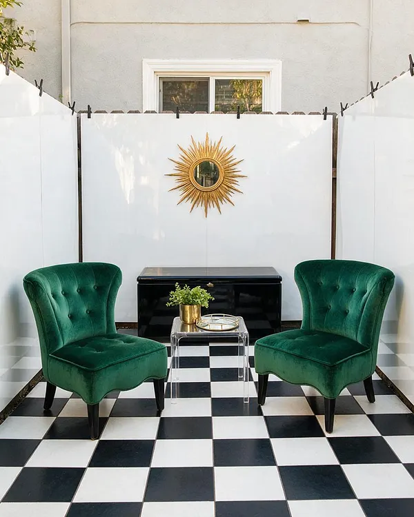A small backyard seating area features two emerald green velvet chairs facing a glossy black cabinet, with a clear acrylic side table between them. White privacy panels surround the space, a gold sunburst mirror hangs on the back wall, and a black-and-white checkerboard floor adds a bold Hollywood Regency look.