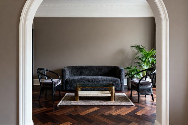 View through a white arched doorway into a calm living room with warm taupe walls and detailed crown molding. Centered is a curved charcoal bouclé sofa flanked by two black rattan armchairs with cushioned seats; a brass coffee table with a glass top and stone shelf sits on a small vintage rug atop walnut herringbone floors, with potted palms to the right and soft daylight filtering from an unseen window.