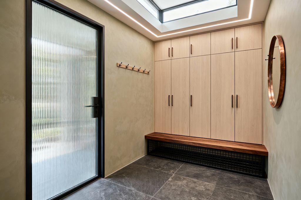 A minimalist mudroom with floor-to-ceiling light wood cabinets featuring slim dark pulls and an integrated walnut bench over a black metal grid for shoe storage. A reeded glass door in a black frame diffuses daylight, while a large skylight with perimeter LED lighting brightens the space. Warm beige plaster-look walls, a simple wood hook rail, a round copper-framed mirror, and charcoal large-format floor tiles create a calm, functional entry.