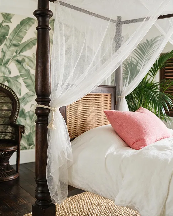 A tropical-style bedroom featuring a dark wood four-poster bed with sheer white canopy drapes, a woven cane headboard, white bedding, and a soft pink accent pillow. Palm-leaf wallpaper and a woven chair in the background reinforce the relaxed, resort-like feel.