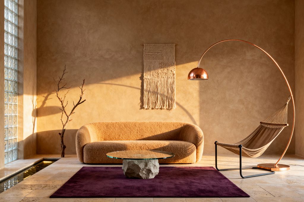 A sunlit modern living room with textured sand-plaster walls and a rounded camel bouclé sofa centered beneath a woven fiber wall hanging. A round glass coffee table rests on a rough stone base atop a deep plum rug. To the right stands an oversized copper arched floor lamp near a tan rope sling chair with a black metal frame. On the left, a glass-block wall filters light toward a narrow indoor water rill set in pale travertine tile flooring.
