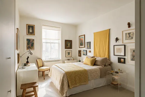 A bright guest bedroom with white walls and a neutral bed layered in cream and tan bedding, accented by a mustard yellow throw and wall hanging. Framed artwork fills the walls, while a small desk, wooden stool, vintage-style chair, and clear side table add an eclectic mix of textures and shapes.