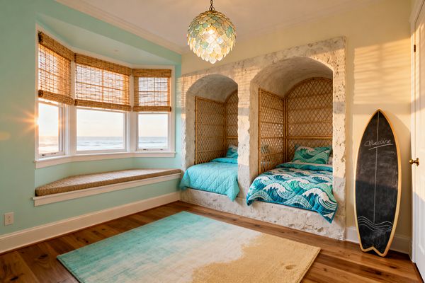 A coastal kids’ room features two twin beds tucked into arched stone niches lined with rattan lattice and dressed in teal wave-pattern quilts. A capiz shell pendant glows above wide-plank wood floors and an aqua-to-sand gradient rug. A bay window with bamboo shades and a woven bench overlooks the ocean at sunset, while a chalkboard surfboard leans by the door. Seafoam walls with white trim create a bright, breezy atmosphere.