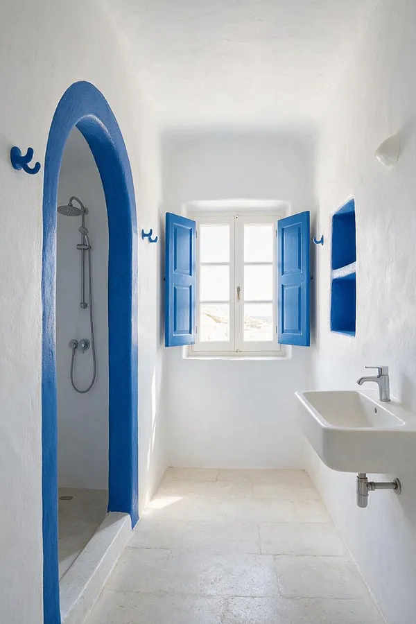 A narrow Mediterranean-style bathroom with white plaster walls, a white stone floor, and vivid cobalt blue trim around an arched shower opening and window shutters. A small wall-mounted sink sits on the right, and bright natural light fills the space through a central window.