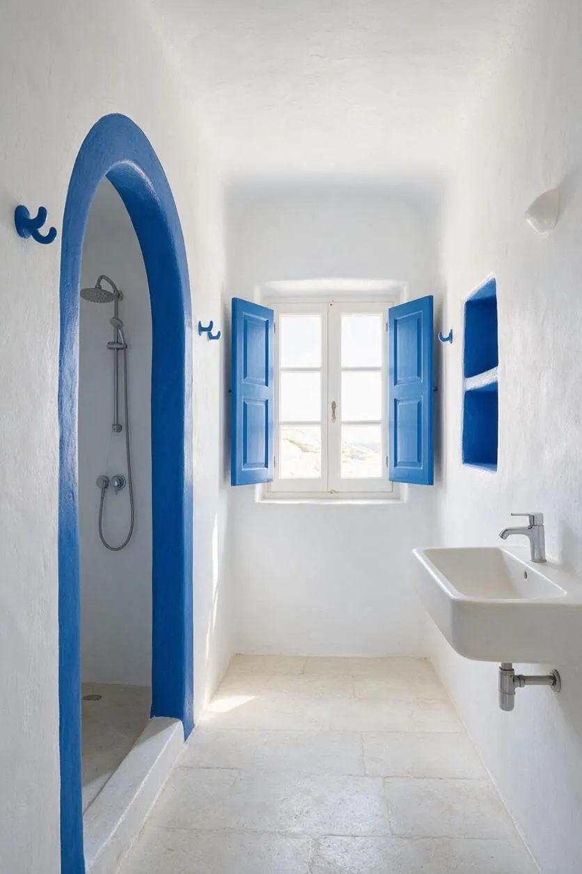 A narrow Mediterranean-style bathroom with white plaster walls, a white stone floor, and vivid cobalt blue trim around an arched shower opening and window shutters. A small wall-mounted sink sits on the right, and bright natural light fills the space through a central window.