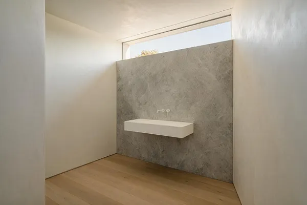 A sparse minimalist powder room with white walls, a light wood floor, and a textured gray stone feature wall at the far end. A simple floating white rectangular sink is mounted on the feature wall beneath a narrow horizontal window that brings in daylight.