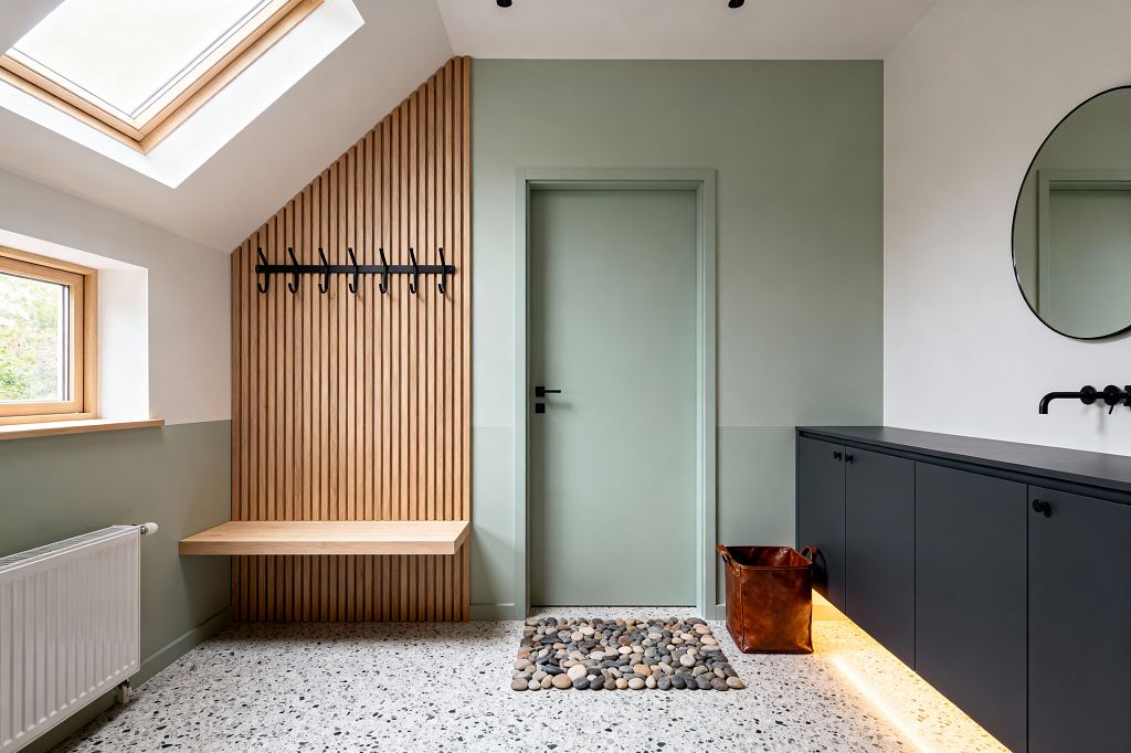 A modern mudroom features two-tone walls in warm white and sage green with a centered sage green door. On the left, a light oak slat wall includes a floating bench and a black metal coat rack. The floor is light terrazzo with gray chips and a rectangular river-stone mat. On the right, a long matte black floating cabinet glows with underlighting, paired with a round mirror and a black wall-mounted faucet. Daylight enters from a skylight and a wood-framed window.