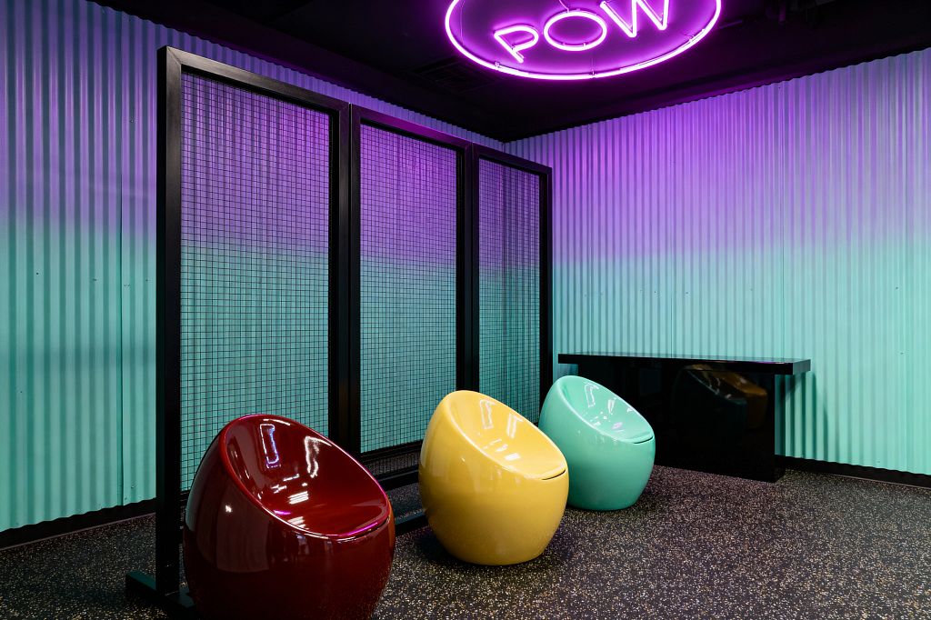 A Pop Art media room with corrugated aqua wall panels washed in violet neon light and a circular ceiling sign reading “POW.” Three glossy pod chairs in cherry red, lemon yellow, and mint green sit on a black speckled floor beside black metal grid partitions and a sleek black console. The space blends industrial textures with playful color and rounded forms for a graphic, lounge-like atmosphere.
