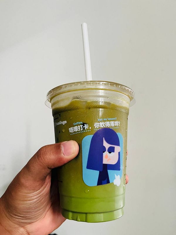 A hand holds a cup of pandan matcha coffee with a playful character sticker on it.