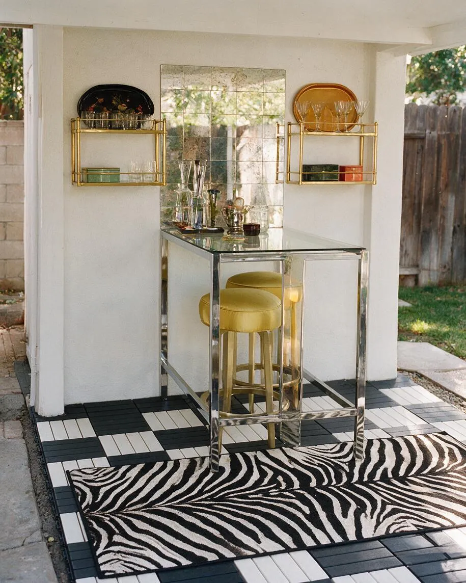 A small backyard bar nook features a mirrored console table with chrome trim and gold stools beneath a mirrored backsplash. Brass wall shelves hold glassware and bar accessories, while a bold black-and-white zebra rug and graphic tile flooring add Hollywood Regency glamour.