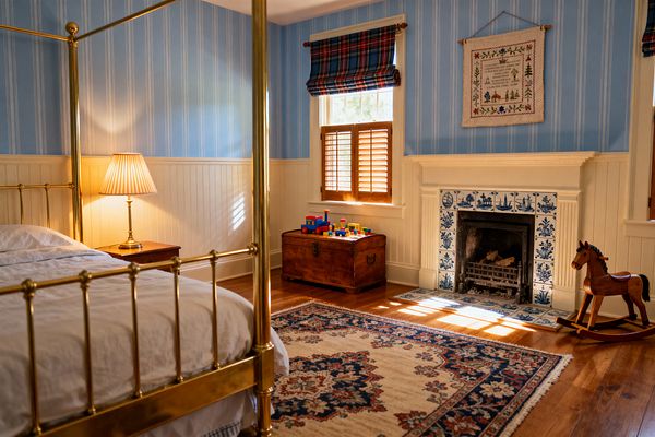 A traditional children’s bedroom with a polished brass four-poster bed and white bedding sits beside a wooden nightstand with a pleated brass lamp. The room features sky-blue striped walls above cream beadboard, honey-toned hardwood floors, a white mantel fireplace faced with blue-and-white Delft-style tiles, a Persian-style rug, a wooden toy chest topped with blocks, plaid Roman shades with wood shutters, and a classic wooden rocking horse in sunlight.