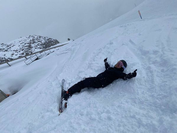A person is lying in the snow on a ski slope, giving a thumbs-up gesture.