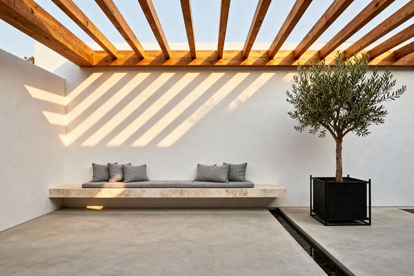 A minimalist patio with white stucco walls and an open wood-beam pergola casting linear shadows. A floating travertine bench runs along the wall, topped with a slim gray cushion and gray throw pillows. At the right, an olive tree stands in a matte black metal planter beside a narrow reflecting water rill, all set on smooth concrete flooring under warm afternoon light.