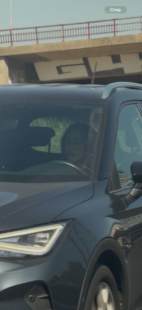 A woman is driving a dark-colored SUV on a highway, visible through the windshield and seemingly using her phone while driving under a bridge.