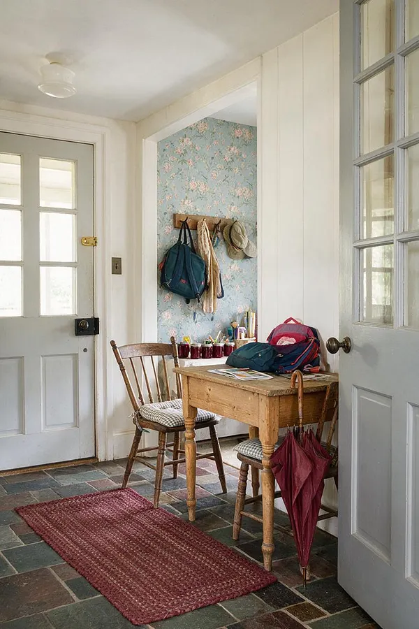 A cottagecore entryway with pale painted doors, a small rustic wooden table, two wooden chairs, and a floral wallpaper alcove with wall hooks holding bags and hats. A woven red runner sits on a stone-tile floor, while soft natural light and aged finishes create a warm, lived-in feel.