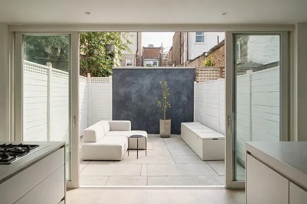 A minimalist backyard courtyard viewed through wide sliding glass doors, featuring white fence panels, pale stone paving, a low white sectional sofa, a small round side table, a long storage bench, and a dark charcoal feature wall with a potted tree. Soft natural light and restrained materials create a calm, modern outdoor retreat.