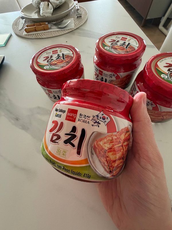 A close-up image of a small glass bowl filled with chopped kimchi placed on a gray textured surface beside a wooden spoon.