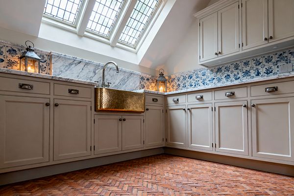 A traditional laundry room features greige inset cabinets with antique nickel hardware, Carrara marble countertops and backsplash, and a hammered brass apron-front sink. A band of blue-and-white Delft-style tiles lines the wall, while a herringbone terracotta brick floor adds warmth. Three leaded-glass skylights bring in natural light, and two candlelit metal lanterns sit on the counter for a soft, ambient glow.