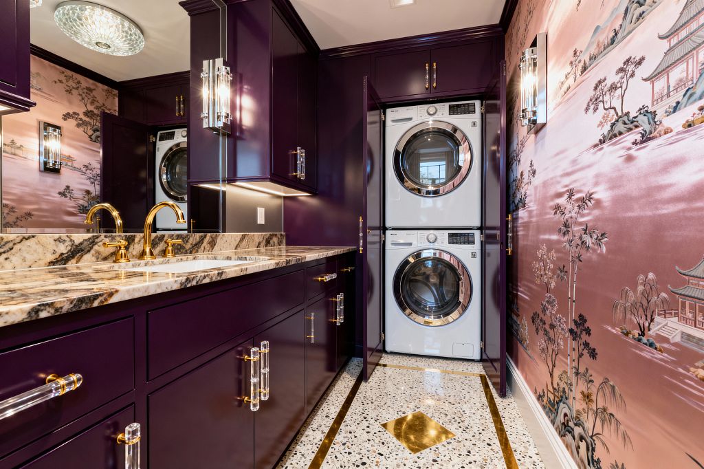 A glamorous laundry room with glossy deep purple cabinetry fitted with clear acrylic and brass pulls, a veined marble countertop with two gold faucets, and a stacked white washer and dryer hidden behind tall cabinet doors. A rose-pink chinoiserie mural covers the opposite wall, while terrazzo flooring with brass inlay runs through the narrow space. Crystal wall sconces, a glass flush-mount ceiling light, and under-cabinet lighting reflect off the lacquered finishes.