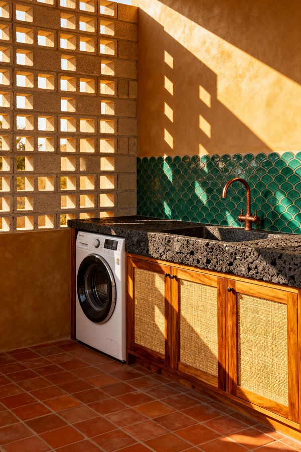 Sunlit tropical laundry room with a perforated concrete breeze-block wall casting geometric shadows over ochre plaster. Along one side, a thick black volcanic-stone countertop with an integrated sink sits above teak cabinets with woven rattan fronts. A copper gooseneck faucet stands before a glossy emerald fish-scale tile backsplash. A white front-loading washer is built into the base, set on terracotta floor tiles.