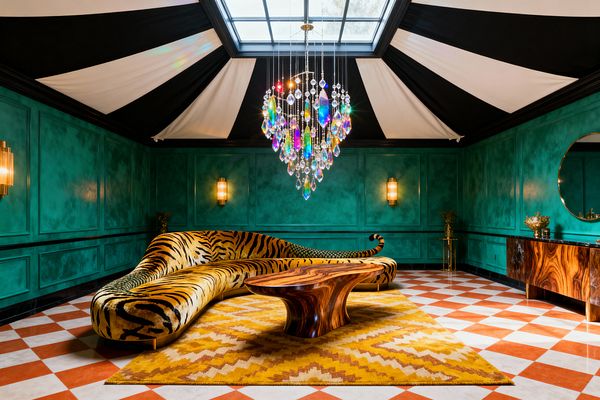 A maximalist living room features emerald green paneled walls and a black-and-white tented ceiling centered by a large skylight. A colorful prism-crystal chandelier hangs above a sculptural wood coffee table and a sweeping curved sofa in tiger-print velvet. A mustard zigzag wool rug rests on a terracotta-and-white checkerboard marble floor. Brass wall sconces glow along the walls, while a richly grained wood credenza and round mirror sit to the right, adding warmth and shine.