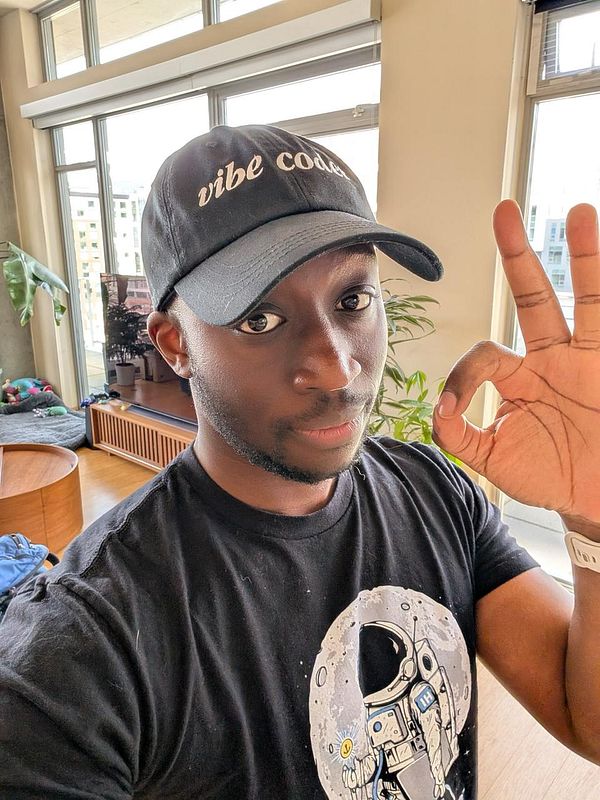 A person wearing a 'vibe coder' hat and an astronaut-themed t-shirt is giving an 'OK' hand gesture.