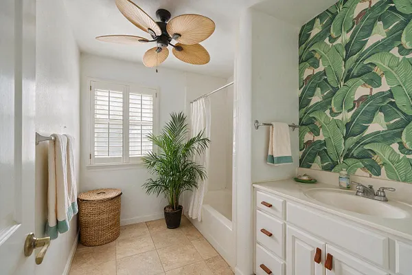 A bright master bathroom with white walls, a tan tile floor, and a vanity on the right topped with a white sink and silver faucet. A large palm-leaf wallpaper accent wall adds tropical color, while a woven basket, potted palm, plantation shutters, and a ceiling fan with leaf-shaped blades complete the relaxed look.