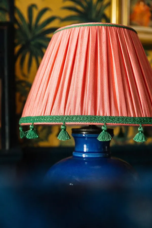 A close-up of a table lamp with a coral pleated fabric shade trimmed in green braid and hanging green tassels. The lamp base is a glossy cobalt blue ceramic or glass form, set against a richly patterned background with tropical motifs and warm yellow tones.