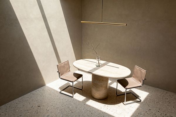 A minimalist dining area lit by a skylight features an oval travertine pedestal table with a clear glass vase holding a single bare branch. Two tan leather sling cantilever chairs with chrome tubular frames face each other. A slim brushed-brass linear pendant hangs above. The space has warm taupe plaster walls and pale speckled terrazzo flooring, with strong sunlight casting crisp shadows that emphasize the clean lines and uncluttered layout.